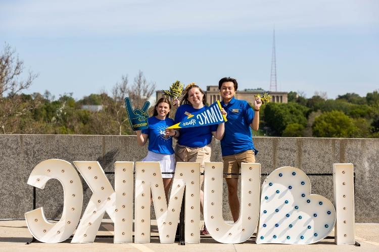 two students wearing blue shirts stand behind a light up sign that reads 'I <3 UMKC'